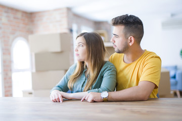 Young couple sitting on the table movinto to new home with carboard boxes behind them looking to side, relax profile pose with natural face with confident smile.