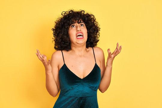 Young arab woman with curly hair wearing elegant dress over isolated yellow background crazy and mad shouting and yelling with aggressive expression and arms raised. Frustration concept.