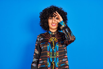 Young arab woman with curly hair wearing colorful shirt over isolated blue background doing ok gesture with hand smiling, eye looking through fingers with happy face.