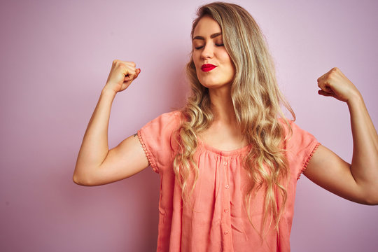 Young beautiful woman wearing t-shirt standing over pink isolated background showing arms muscles smiling proud. Fitness concept.