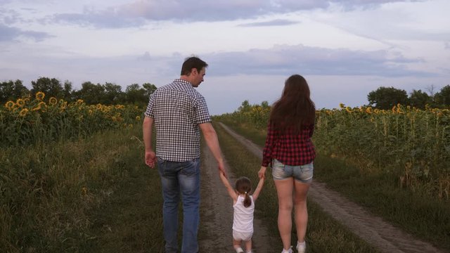 little daughter jumping holding hands mom and dad. Family with small child walks along road and laughs next to field of sunflowers. Mom, dad and daughter are resting together outside city in nature.