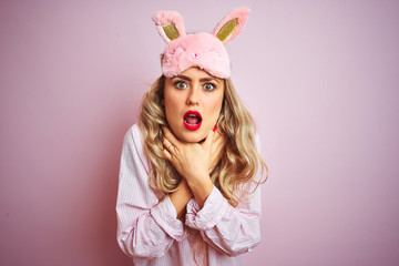 Young beautiful woman wearing pajama and sleep mask over pink isolated background shouting and suffocate because painful strangle. Health problem. Asphyxiate and suicide concept.