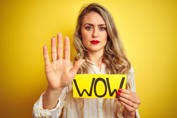 Beautiful woman holding amazed wow surprise banner over isolated yellow background with open hand doing stop sign with serious and confident expression, defense gesture