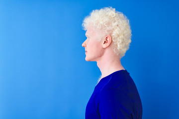 Young albino blond man wearing casual t-shirt standing over isolated blue background looking to side, relax profile pose with natural face with confident smile.