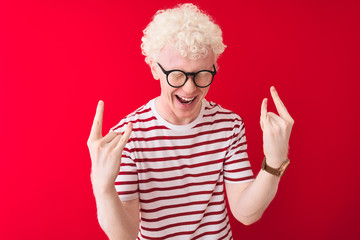 Young albino blond man wearing striped t-shirt and glasses over isolated red background shouting with crazy expression doing rock symbol with hands up. Music star. Heavy concept.