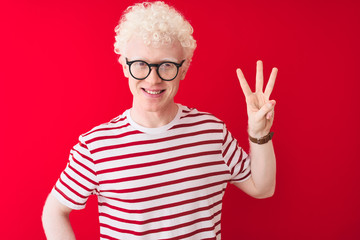 Young albino blond man wearing striped t-shirt and glasses over isolated red background showing and pointing up with fingers number three while smiling confident and happy.