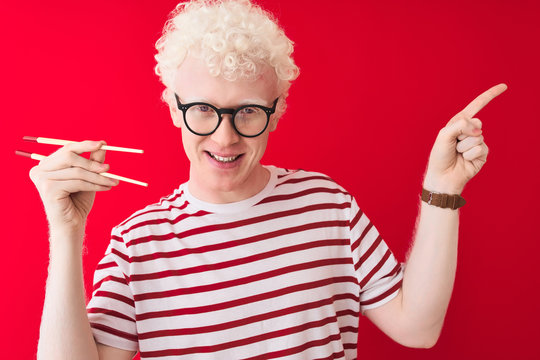 Young albino blond man holding chopsticks standing over isolated white background very happy pointing with hand and finger to the side