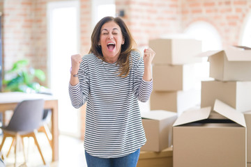 Middle age woman moving to a new house arround cardboard boxes celebrating surprised and amazed for success with arms raised and open eyes. Winner concept.