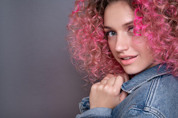 Naklejka premium Portrait of beautiful woman with pink curly colored hairs in blue jeans jacket. Glitter makeup. 90s style. Ombre. Isolated on grey