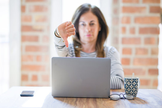 Middle age senior woman sitting at the table at home working using computer laptop with angry face, negative sign showing dislike with thumbs down, rejection concept