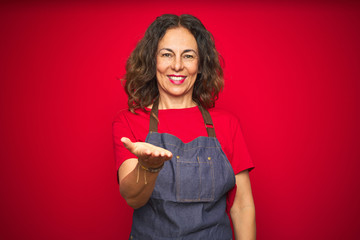 Middle age senior woman wearing apron uniform over red isolated background smiling cheerful offering palm hand giving assistance and acceptance.