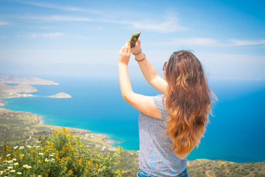Young woman making selfie on a beautiful background of aegean sea and mountains. Good-looking young woman with brown wavy hair taking picture of herself.