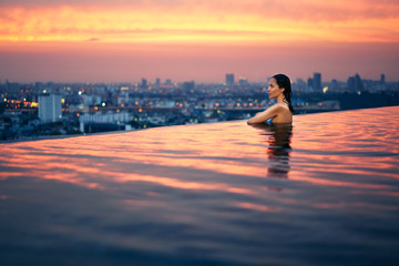 Young woman relax in swimming pool on roof top during amazing sunset and enjoy cityscape