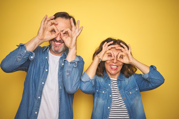 Beautiful middle age couple together wearing denim shirt over isolated yellow background doing ok gesture like binoculars sticking tongue out, eyes looking through fingers. Crazy expression.