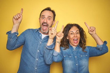 Beautiful middle age couple together standing over isolated yellow background smiling amazed and surprised and pointing up with fingers and raised arms.