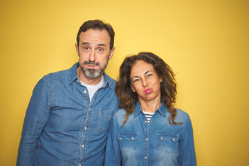Fototapeta premium Beautiful middle age couple together standing over isolated yellow background puffing cheeks with funny face. Mouth inflated with air, crazy expression.