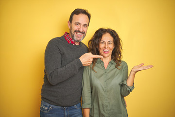 Beautiful middle age couple over isolated yellow background amazed and smiling to the camera while presenting with hand and pointing with finger.
