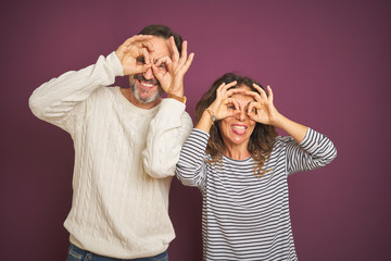 Beautiful middle age couple wearing winter sweater over isolated purple background doing ok gesture like binoculars sticking tongue out, eyes looking through fingers. Crazy expression.