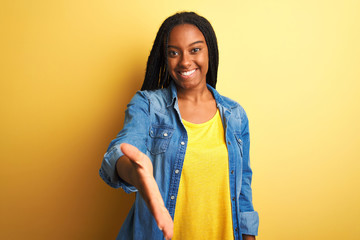 Young african american woman wearing denim shirt standing over isolated yellow background smiling friendly offering handshake as greeting and welcoming. Successful business.