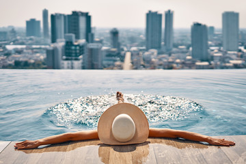 Back view of young woman in hat relaxing in swimming pool on the roof top of hotel