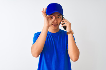Chinese deliveryman wearing cap talking on the smartphone over isolated white background with happy face smiling doing ok sign with hand on eye looking through fingers