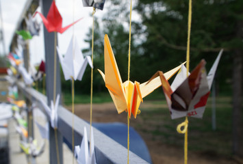 Origami multicolored paper cranes tied to threads on the fence of a city park. Shallow depth of field