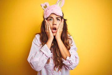 Young woman wearing pajama and sleep mask standing over yellow isolated background afraid and shocked, surprise and amazed expression with hands on face