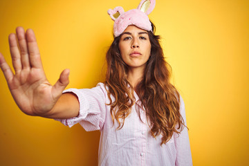 Young woman wearing pajama and sleep mask standing over yellow isolated background doing stop sing with palm of the hand. Warning expression with negative and serious gesture on the face.