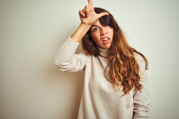 Fototapeta premium Young beautiful woman wearing winter sweater standing over white isolated background making fun of people with fingers on forehead doing loser gesture mocking and insulting.