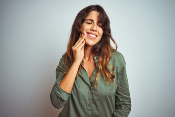 Young beautiful woman wearing green shirt standing over grey isolated background touching mouth with hand with painful expression because of toothache or dental illness on teeth. Dentist concept.