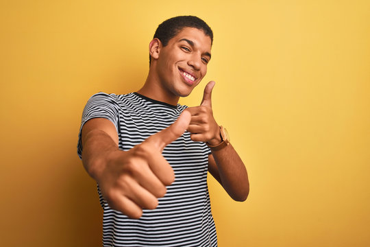 Young handsome arab man wearing navy striped t-shirt over isolated yellow background approving doing positive gesture with hand, thumbs up smiling and happy for success. Winner gesture.