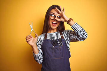 Young beautiful hairdresser woman holding scissors standing over isolated yellow background with happy face smiling doing ok sign with hand on eye looking through fingers