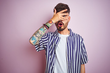 Young man with tattoo wearing striped shirt standing over isolated pink background peeking in shock covering face and eyes with hand, looking through fingers with embarrassed expression.