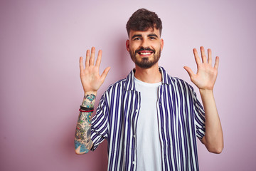 Fototapeta premium Young man with tattoo wearing striped shirt standing over isolated pink background showing and pointing up with fingers number ten while smiling confident and happy.