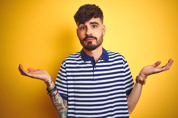 Young man with tattoo wearing striped polo standing over isolated yellow background clueless and...