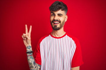 Young man with tattoo wearing striped t-shirt standing over isolated red background smiling with happy face winking at the camera doing victory sign. Number two.