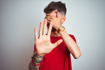 Young man with tattoo wearing red t-shirt standing over isolated white background covering eyes with hands and doing stop gesture with sad and fear expression. Embarrassed and negative concept.