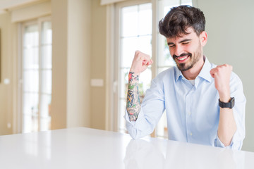 Fototapeta premium Young businesss man sitting on white table very happy and excited doing winner gesture with arms raised, smiling and screaming for success. Celebration concept.