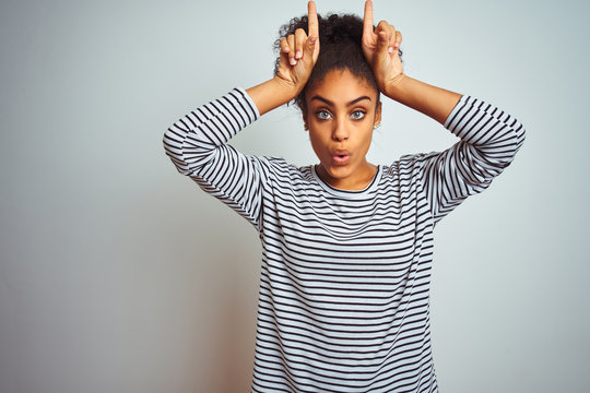 African american woman wearing navy striped t-shirt standing over isolated white background doing funny gesture with finger over head as bull horns