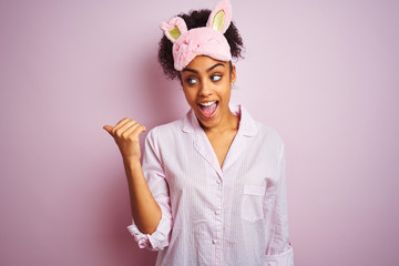 Young african american woman wearing pajama and mask over isolated pink background smiling with happy face looking and pointing to the side with thumb up.