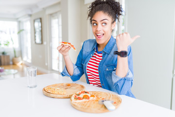 Young african american woman eating two homemade cheese pizzas pointing and showing with thumb up to the side with happy face smiling