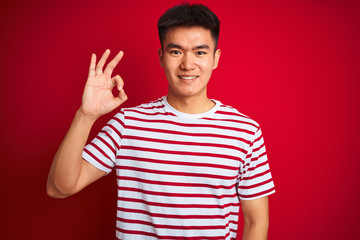 Naklejka premium Young asian chinese man wearing striped t-shirt standing over isolated red background smiling positive doing ok sign with hand and fingers. Successful expression.