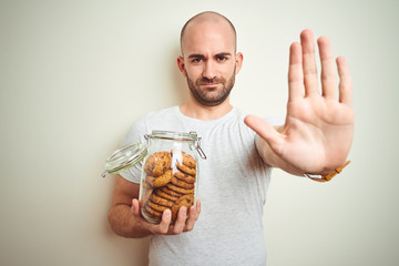 Young man holding jar of chocolate chips cookies over isolated background with open hand doing stop sign with serious and confident expression, defense gesture