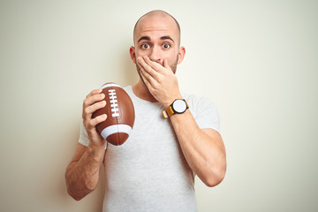 Young man holding rugby american football ball over isolated background cover mouth with hand...