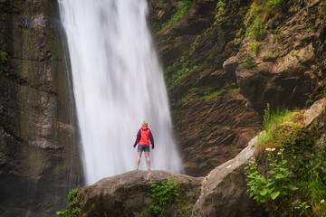 Hiking woman in red jacket stay opposite big waterfall in mountains.