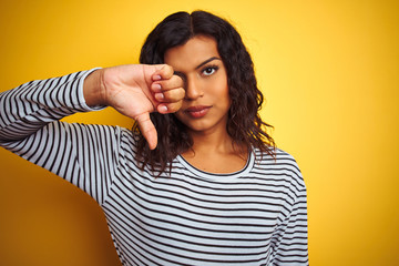 Transsexual transgender woman wearing striped t-shirt over isolated yellow background with angry face, negative sign showing dislike with thumbs down, rejection concept