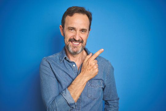 Handsome middle age senior man with grey hair over isolated blue background cheerful with a smile on face pointing with hand and finger up to the side with happy and natural expression