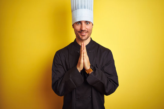 Young chef man wearing uniform and hat standing over isolated yellow background praying with hands together asking for forgiveness smiling confident.