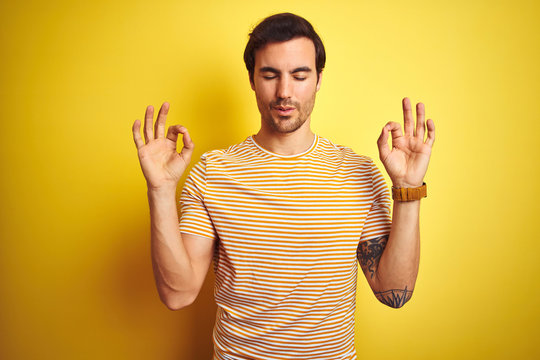Young handsome man with tattoo wearing striped t-shirt over isolated yellow background relax and smiling with eyes closed doing meditation gesture with fingers. Yoga concept.