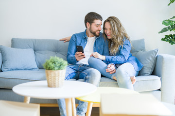 Young couple in love hugging sitting on the sofa using smartphone very happy moving to a new home